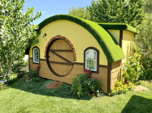 A whimsical yellow hobbit-style house with a rounded green roof, surrounded by colorful flowers and greenery.